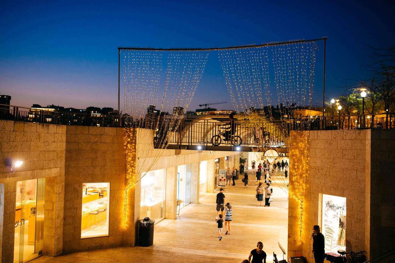 Mamilla Mall open-air shopping area at dusk with illuminated stone facades and decorative light installations overhead.