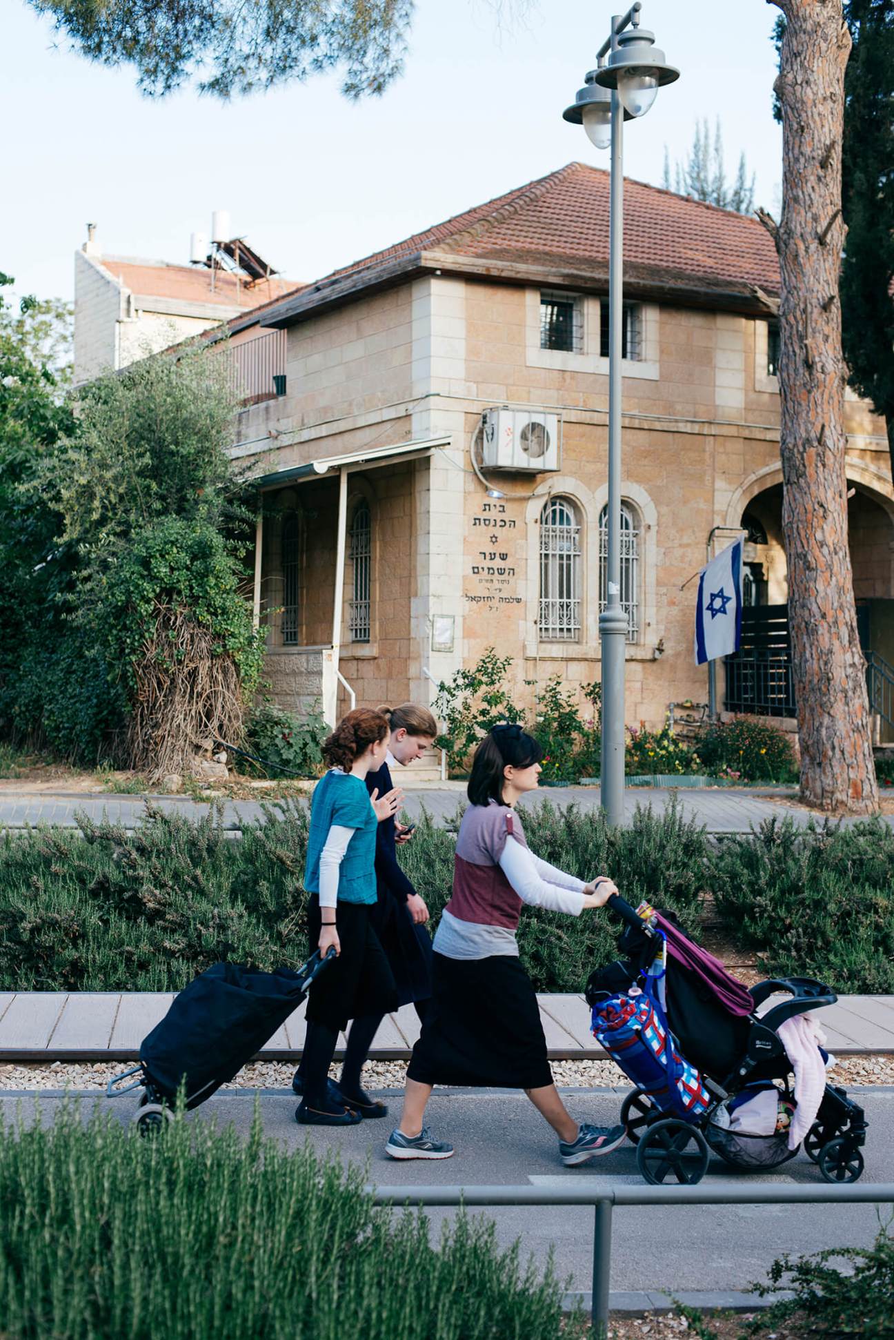 Two women and a child with stroller walk past a stone building with Israeli flag in German Colony, Jerusalem.