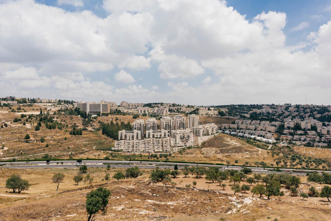 Aerial view of Jerusalem showing residential buildings on hillsides with a highway, olive trees, and scattered development across the landscape.