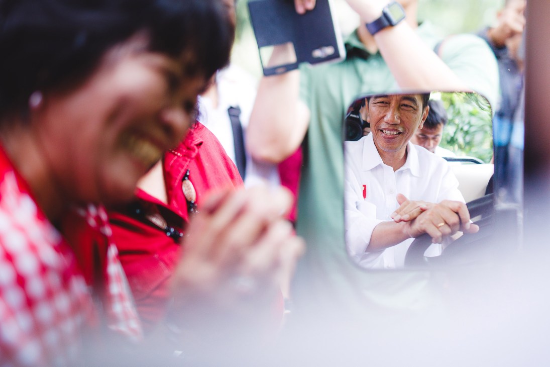 Indonesian President Joko Widodo smiles while sitting in an open vehicle surrounded by crowds of people taking photos and selfies outside the presidential palace.
