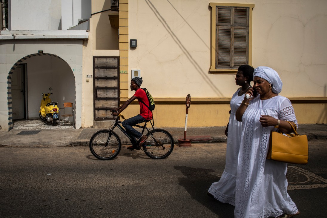 Two women in white traditional dress walk past a cyclist in red on a street in Dakar's old town