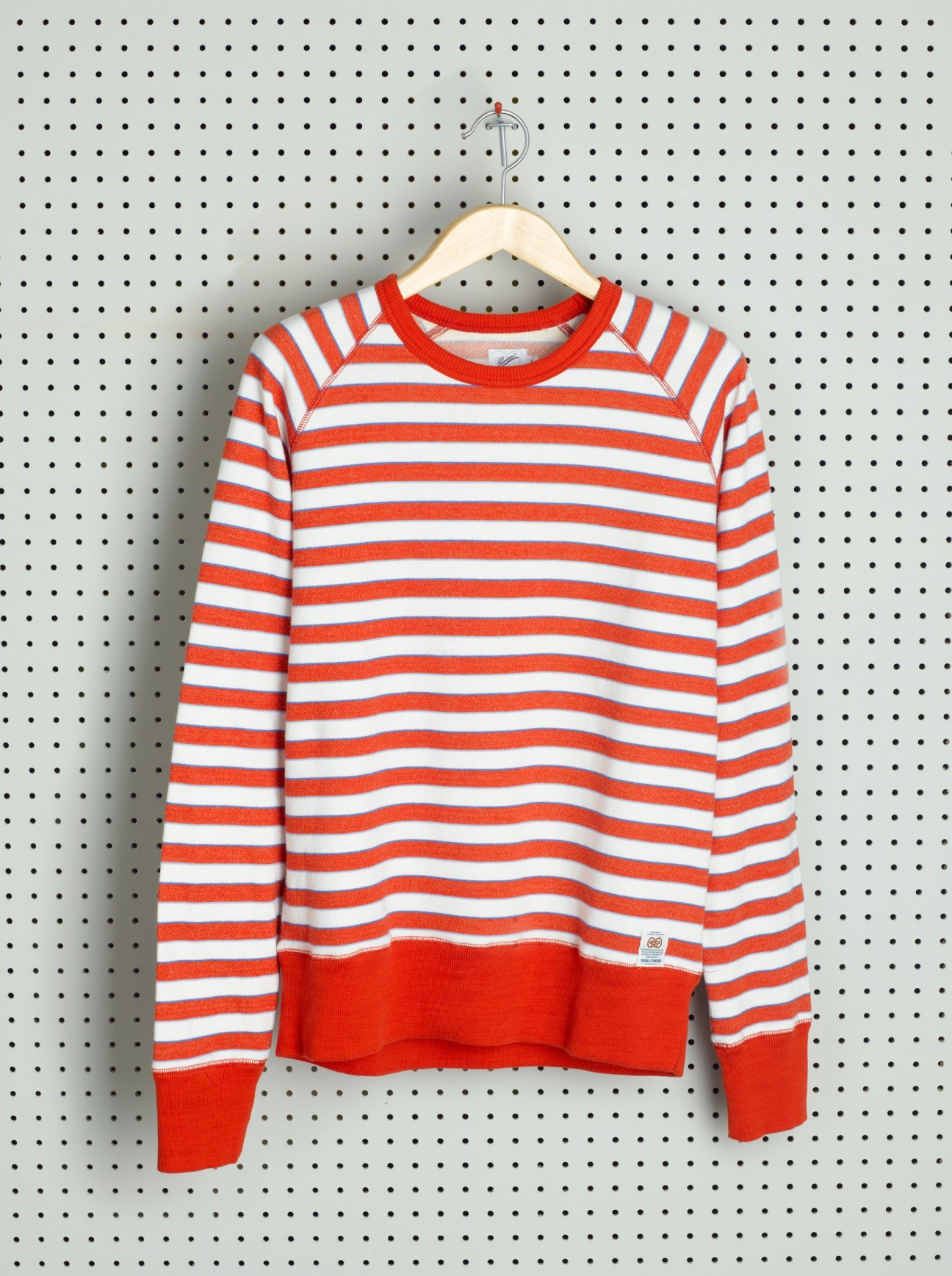Red and white striped long-sleeve jumper on wooden hanger against pegboard background.