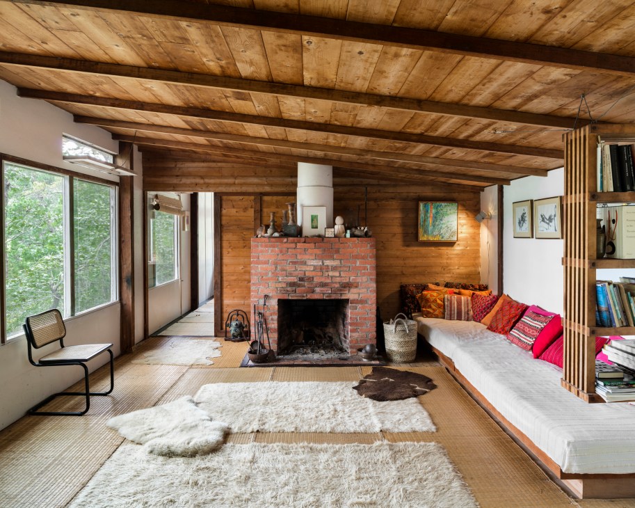 Living room at Kepes House designed by Marcel Breuer, featuring exposed wooden beams, brick fireplace, and large windows overlooking the pond.
