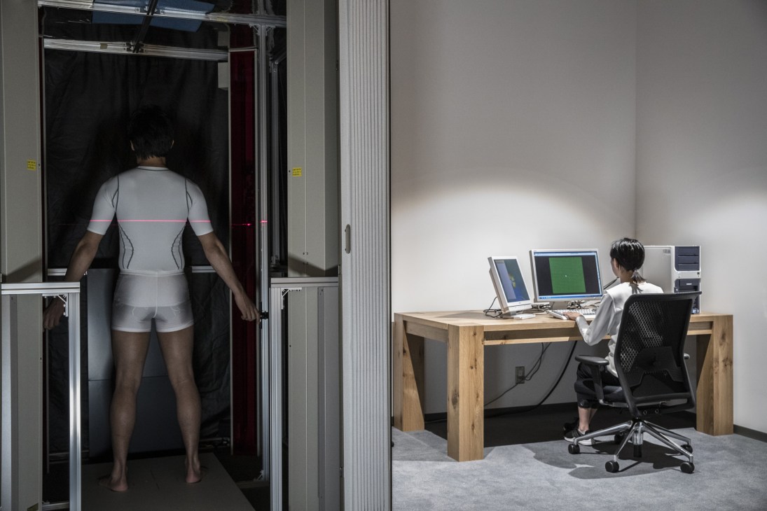 Person in light-colored athletic wear stands in body scanner chamber while technician monitors fitting on computer
