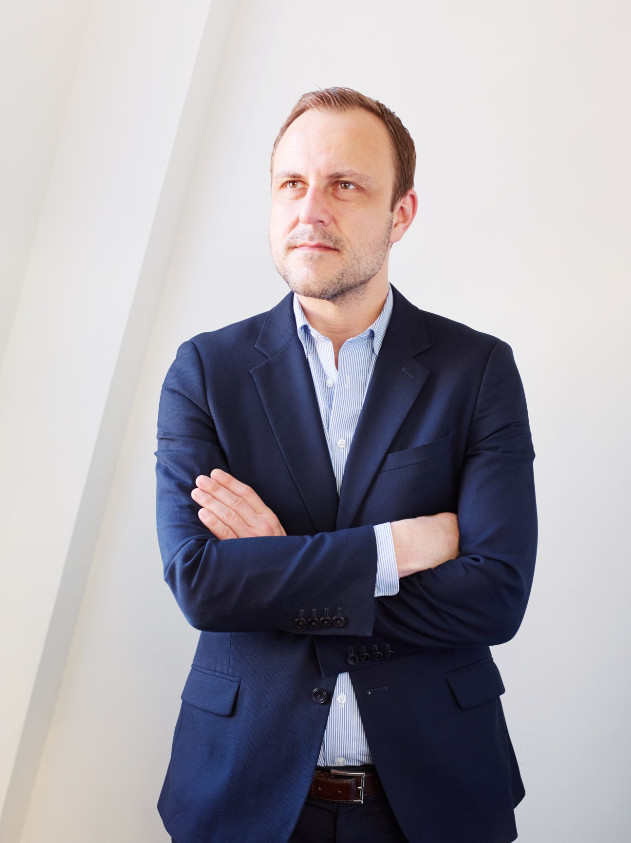 Portrait of Peter Neumann, a man in a dark blazer and striped shirt standing with arms crossed against a white background.