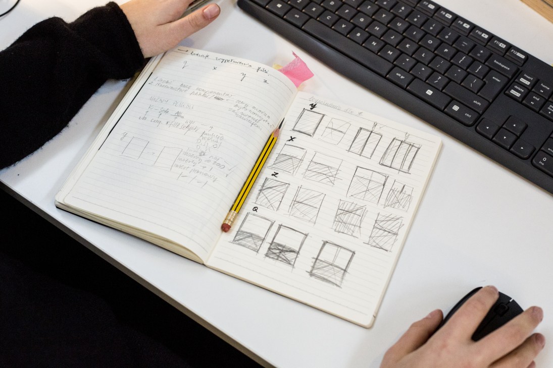 Designer's hands holding a notebook with furniture sketches and design iterations next to a computer keyboard.