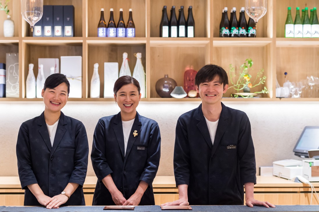 Three smiling staff members in navy blazers stand behind a counter at Imadeya liquor store in Tokyo.