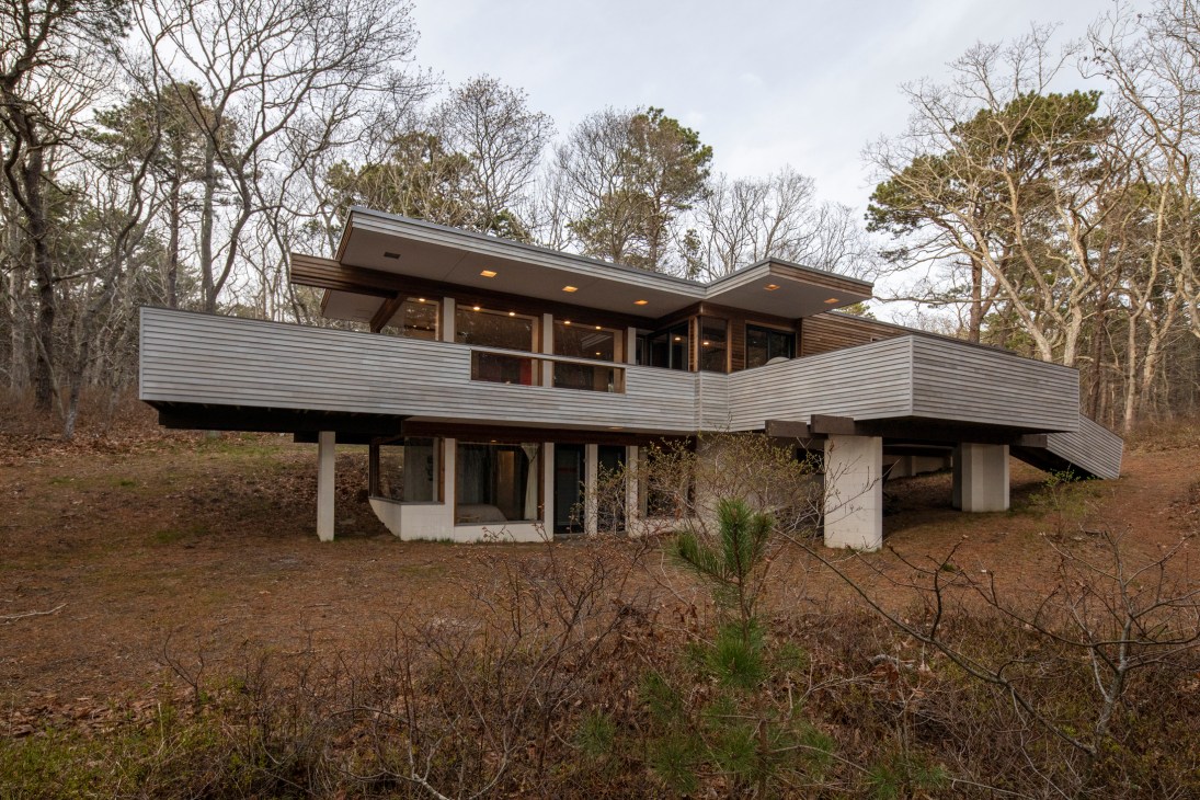 Modernist house with horizontal metal railings elevated on concrete pillars surrounded by bare trees and woodland.