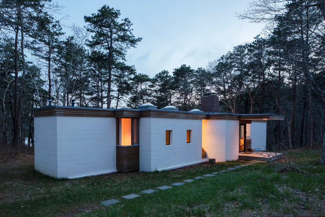 White modernist house with curved walls and dark trim nestled in pine forest at dusk.