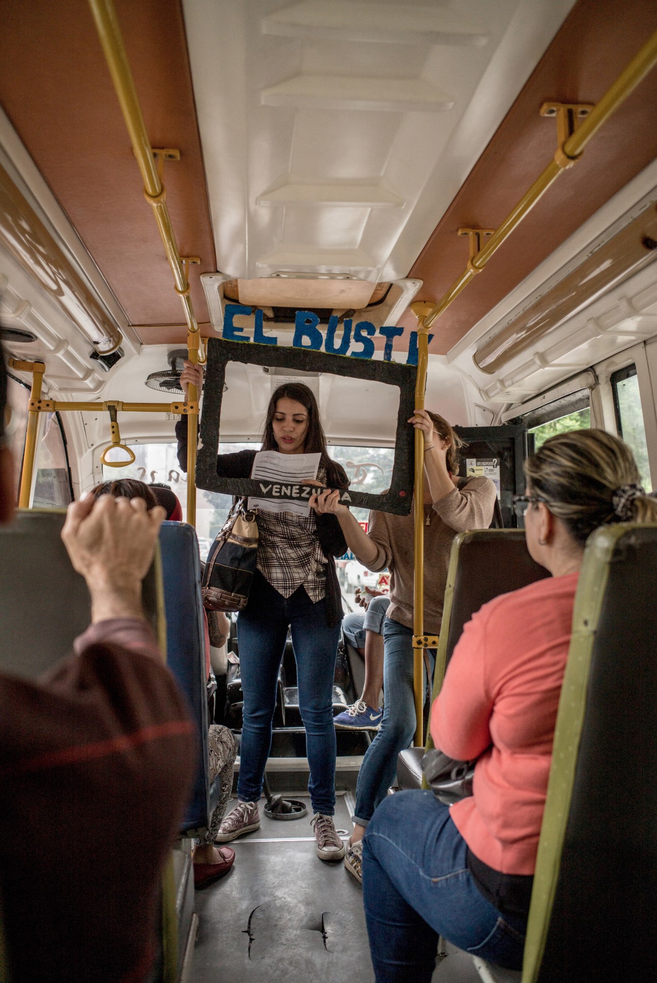 Woman holding El Bus TV sign while standing on public bus with passengers seated around her
