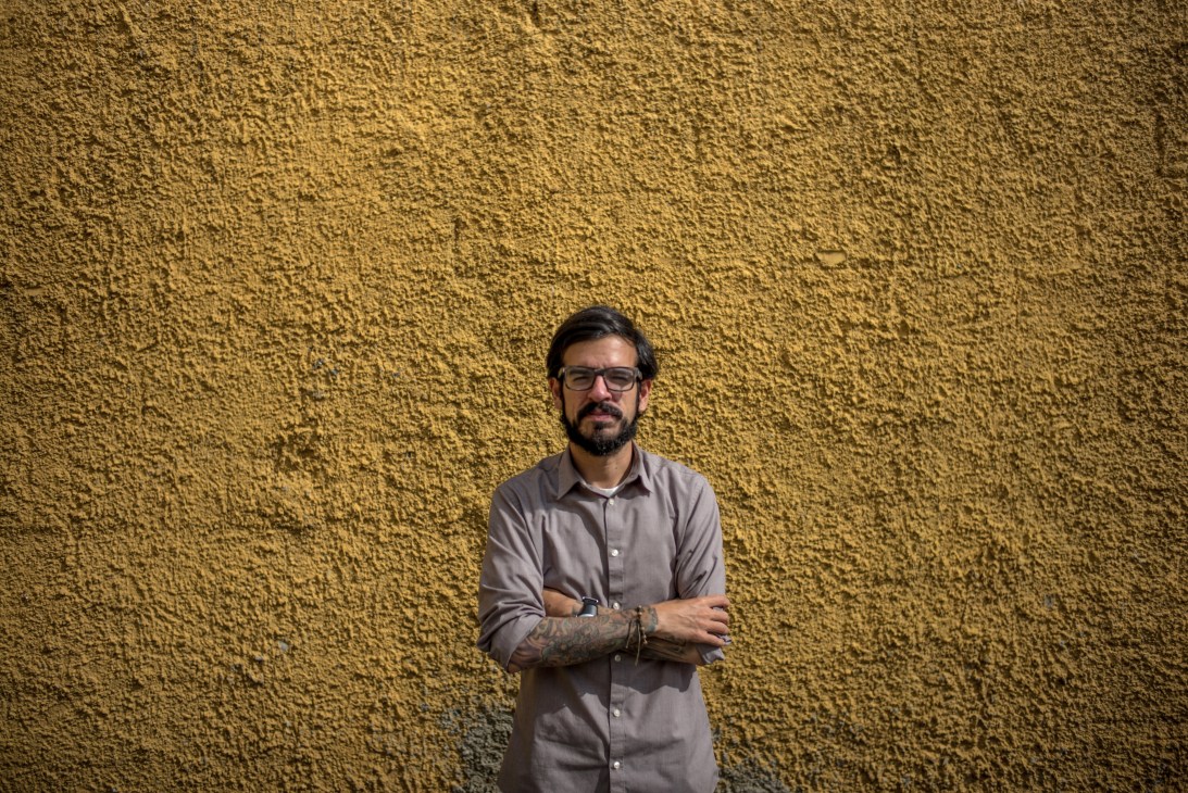 Man with glasses and beard in light colored shirt with crossed arms against textured yellow wall