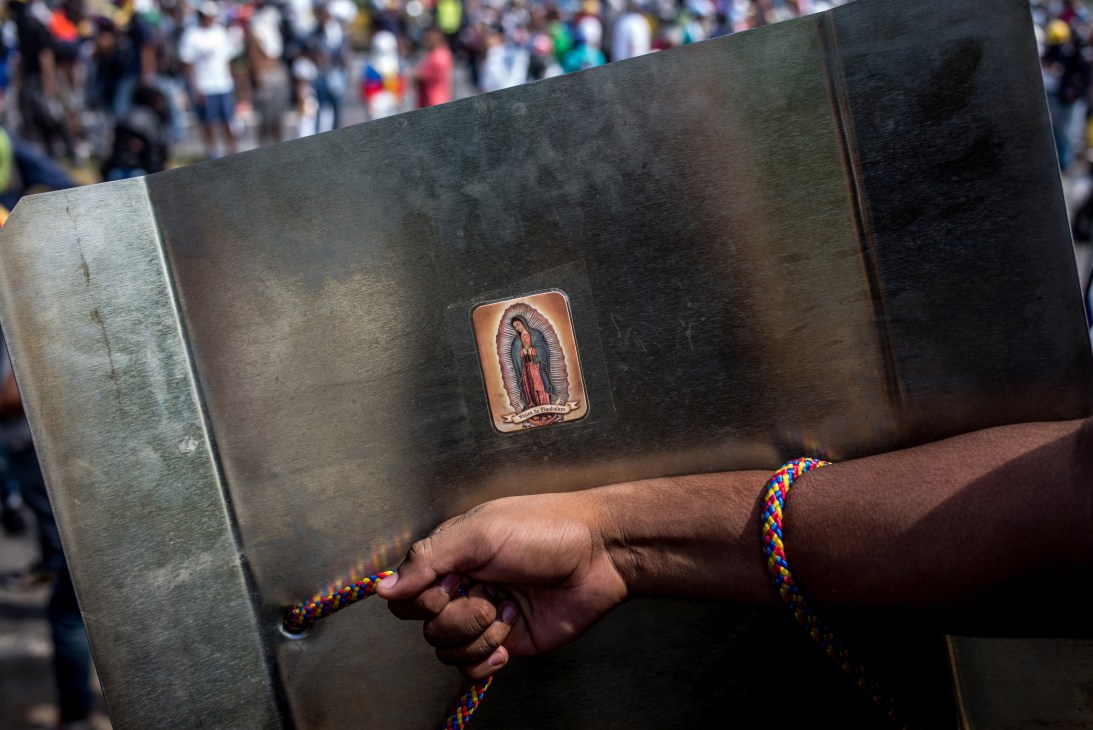 Person holding makeshift shield with Virgin Mary sticker and colorful braided rope strap during protest