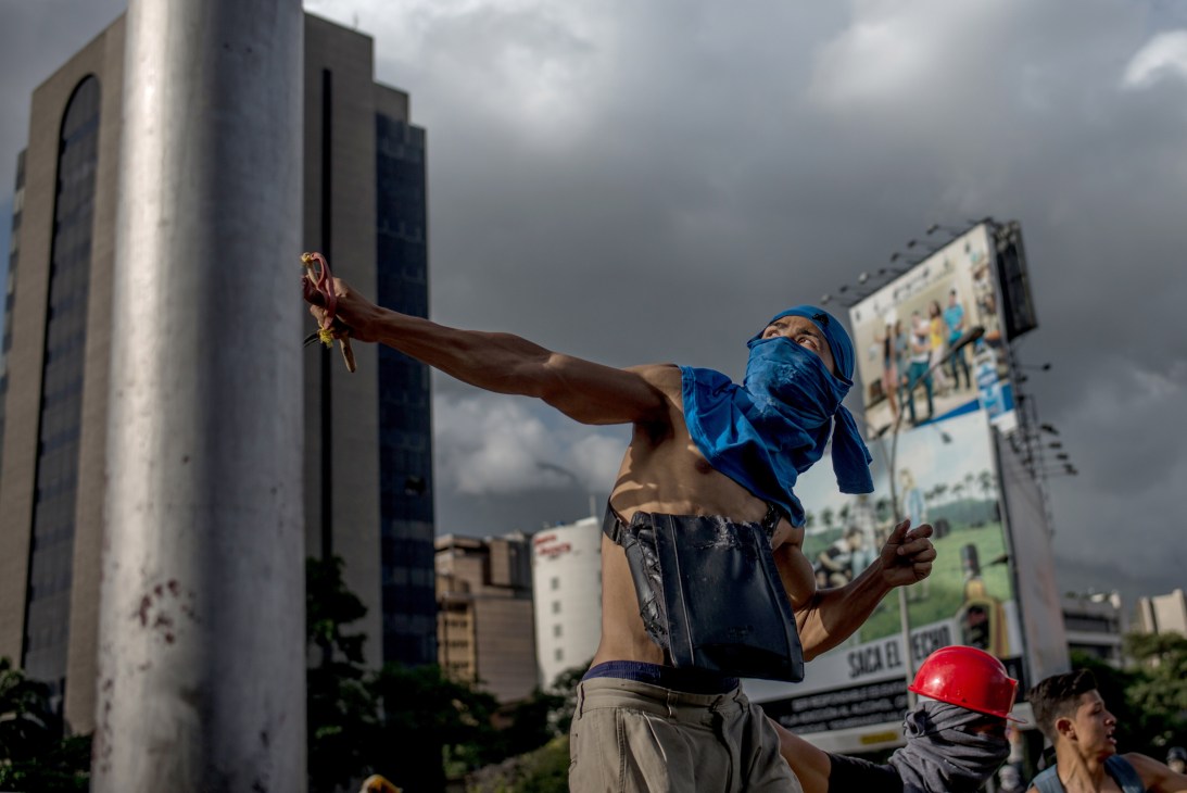 Protester with blue face covering using homemade slingshot in urban setting with buildings in background