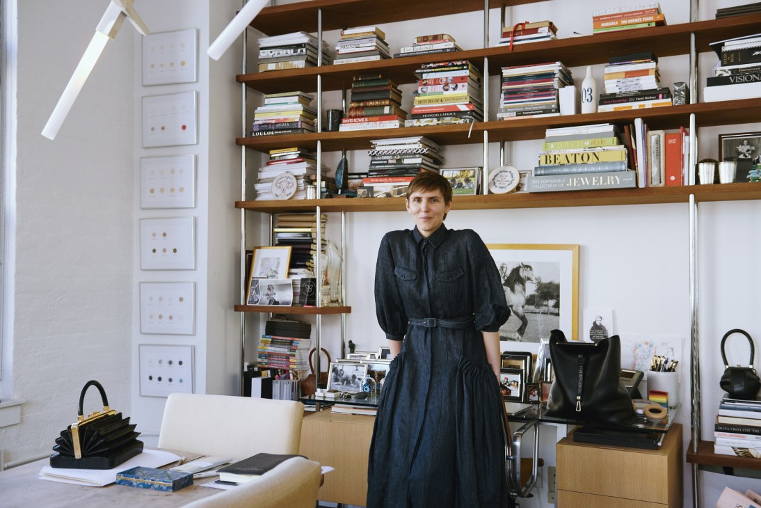 Gabriela Hearst standing in her sun-filled showroom office with wooden shelves of books and design materials behind her