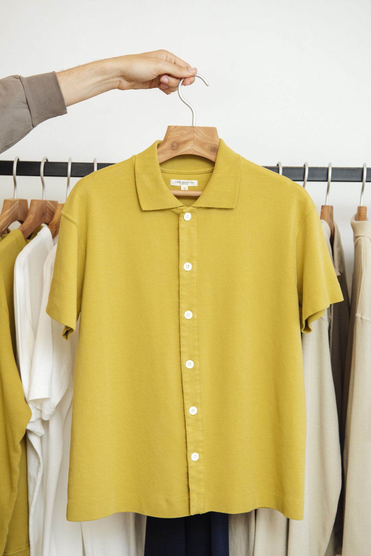 Yellow button-down shirt on wooden hanger held by hand, with neutral clothing on rack behind.