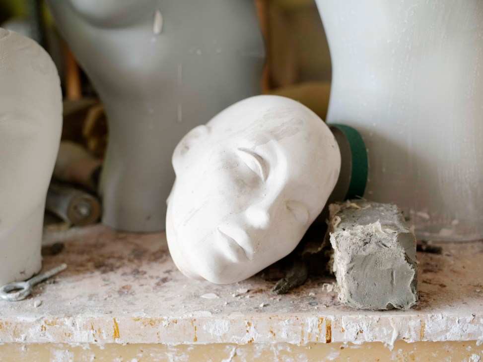 White plaster mannequin head with closed eyes and defined lips among other unpainted head forms in La Rosa's workshop.