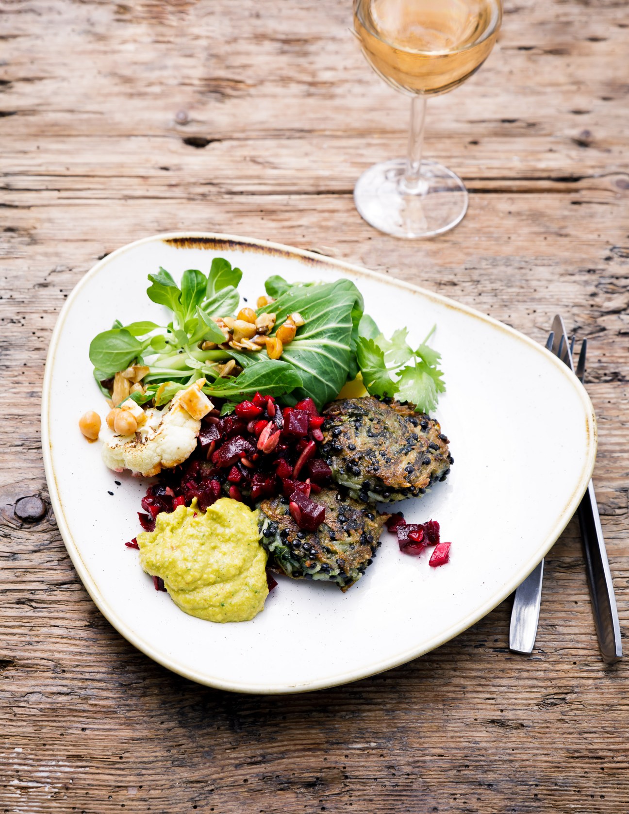 Lentil patties served on white plate with mixed greens, beetroot salad, green puree and wine glass