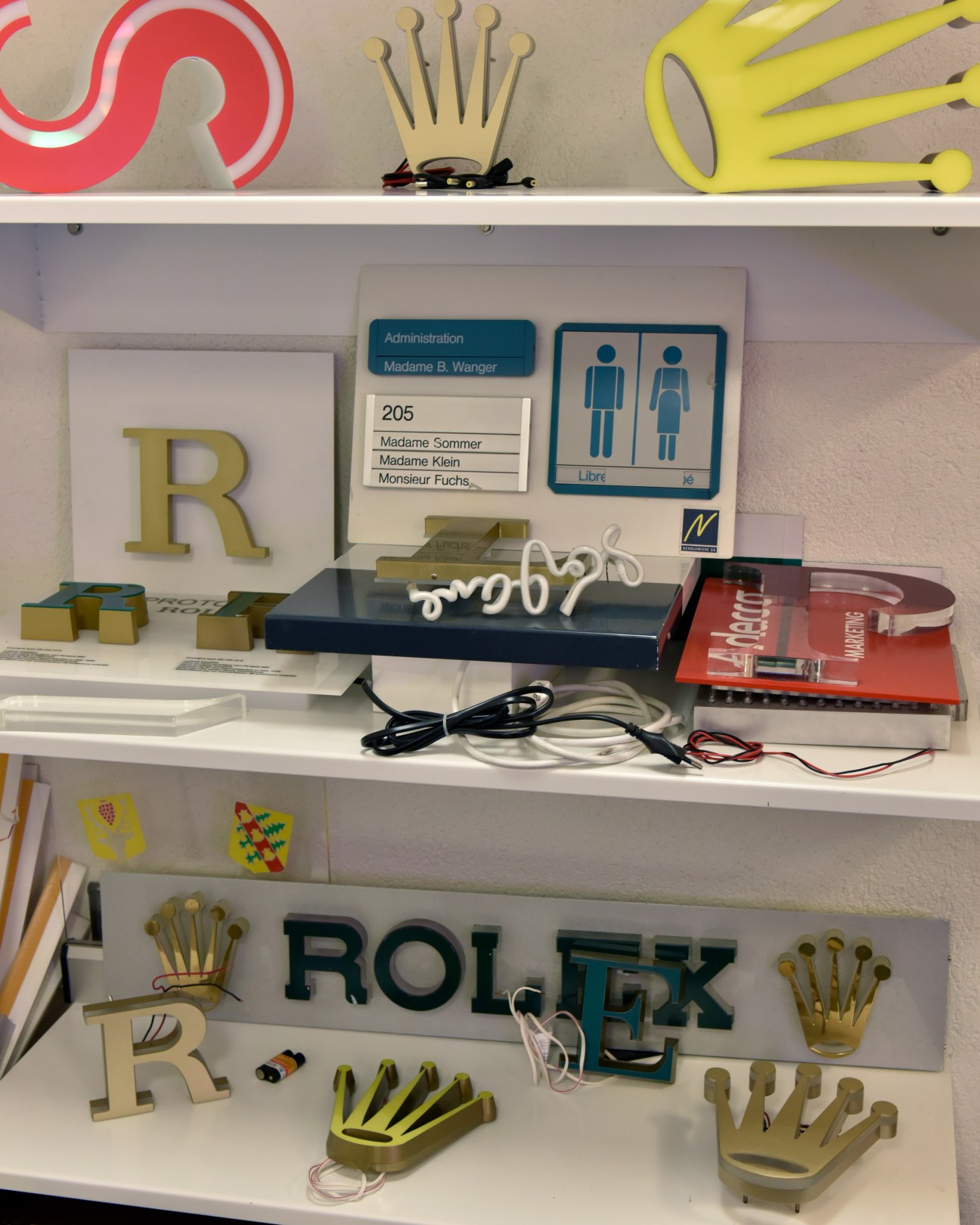 Shelves displaying prototype Rolex signs, letters, and crown logos in various colors and materials