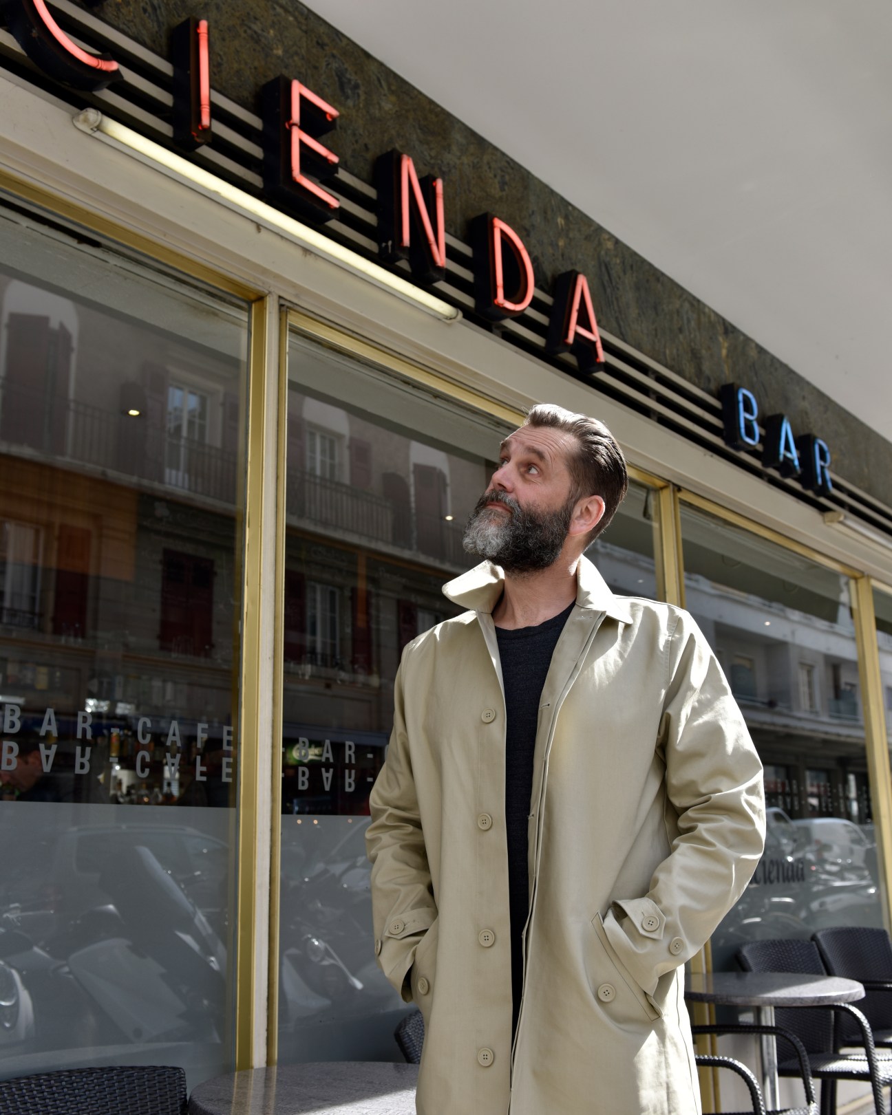 Olivier Walker stands outside a cafe looking up, wearing a beige coat over a dark shirt