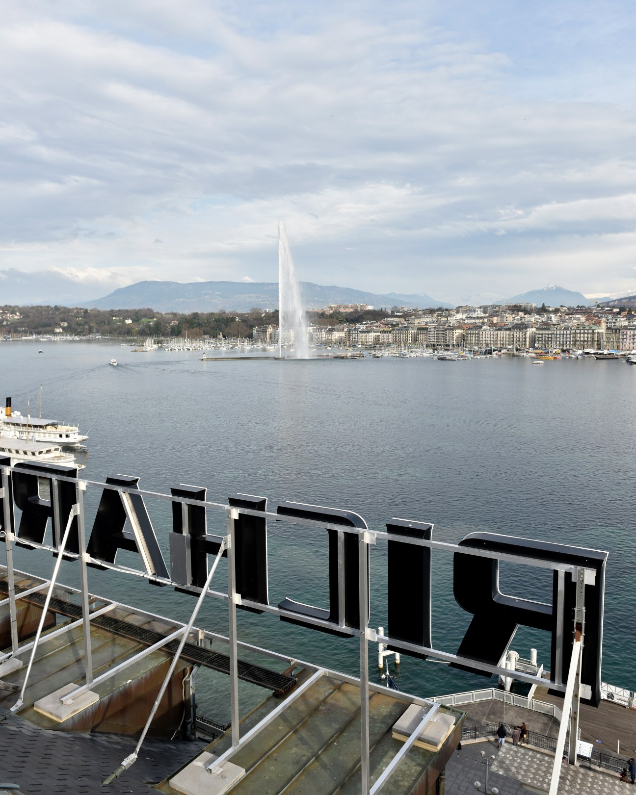 View from boat deck showing Geneva's lakefront with fountain and neon signs on buildings in distance