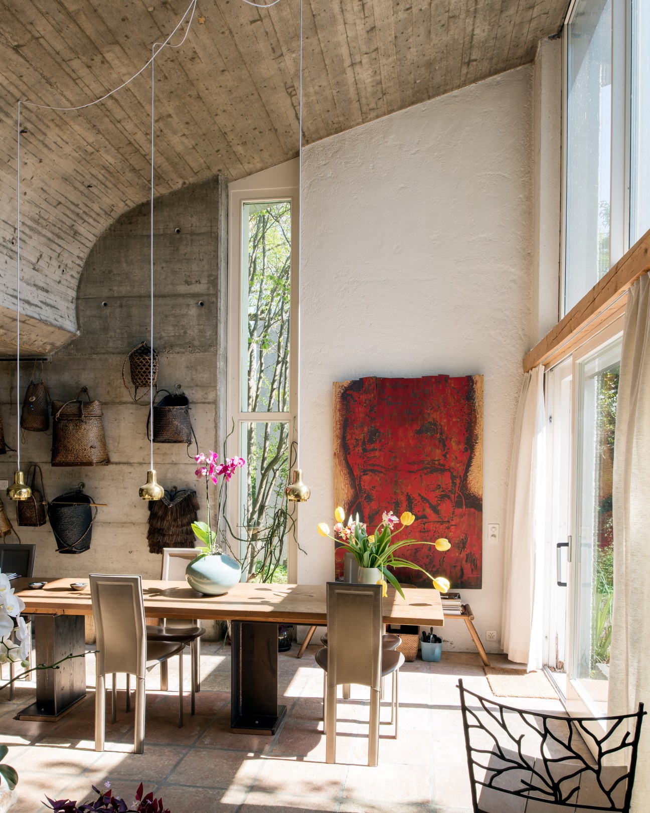 Open-plan dining area with exposed concrete curved ceiling, pendant lamps, wooden table, and red abstract artwork on white wall.