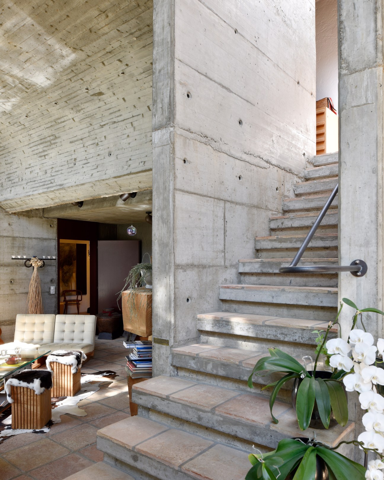 Concrete staircase with metal handrail in a modernist home interior with exposed concrete walls and living area.