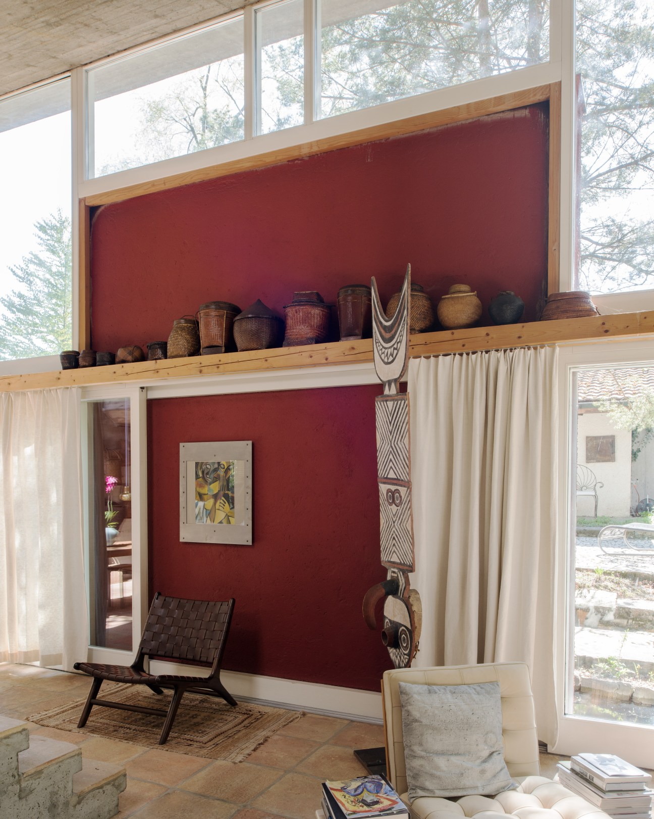 Living room with ochre-red accent wall displaying African sculptures and baskets, modernist furniture, and large windows overlooking trees.
