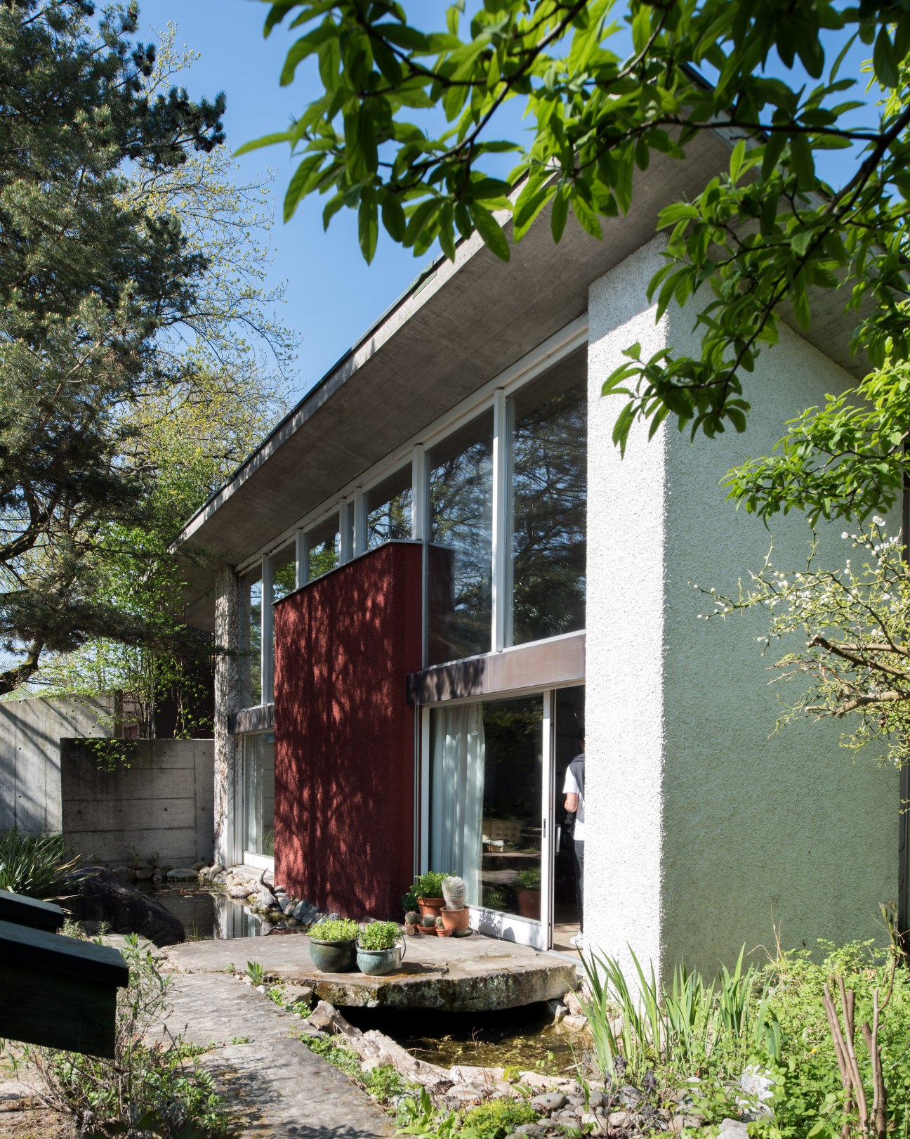Modernist concrete house exterior with ochre-painted wall, large glass doors, and surrounding vegetation.