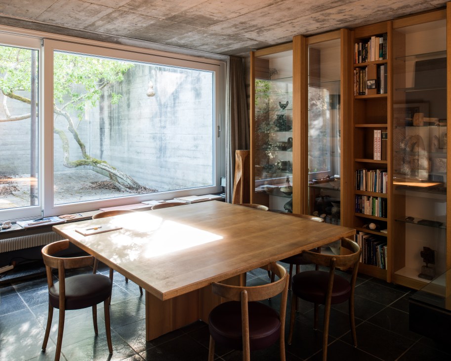 Modernist architecture studio with wooden dining table, chairs, built-in shelving, large windows overlooking a concrete courtyard wall and natural landscape.