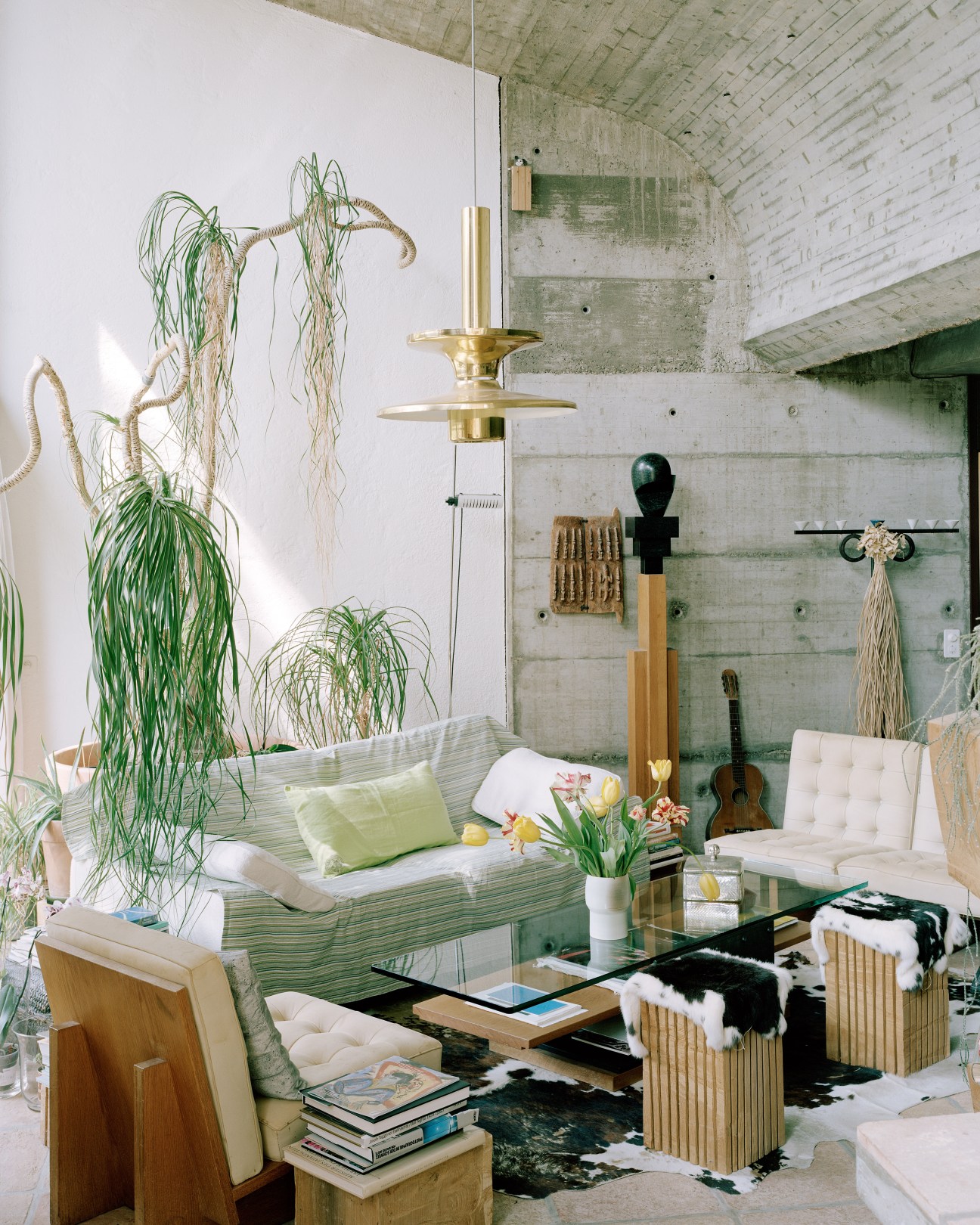 Living room interior with exposed concrete walls, Eduard Neuenschwander-designed furniture, brass lamp, and abundant potted plants.