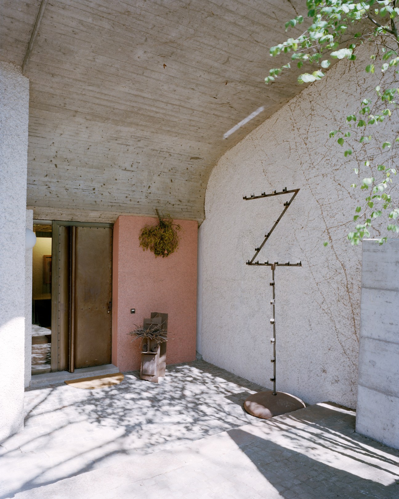 Modern concrete entranceway with sculptural metal numerals, ochre-painted wall, and climbing vines under a textured ceiling.