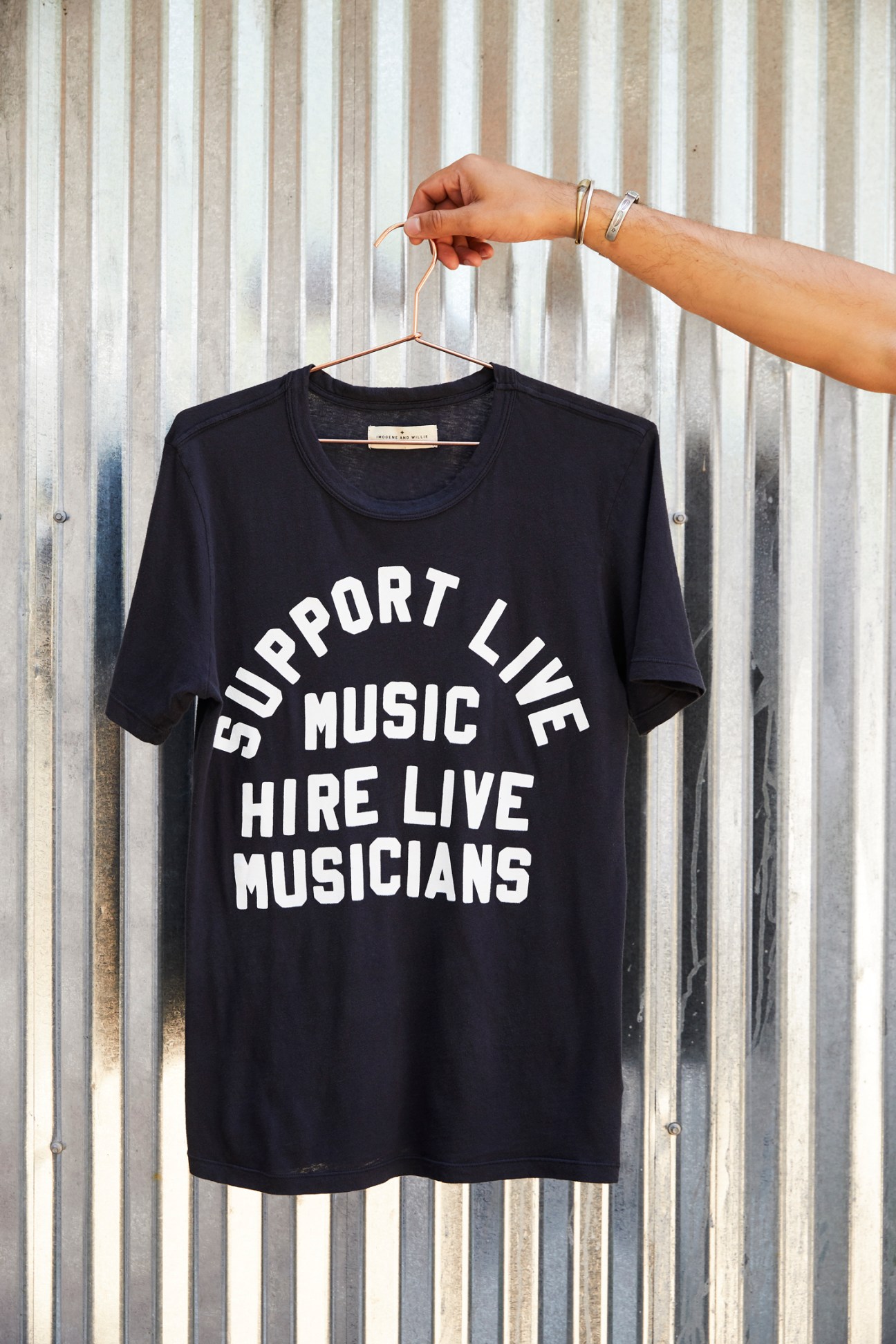 Black t-shirt with white text reading 'Support Live Music Hire Live Musicians' hanging on a wooden hanger against a corrugated metal background.