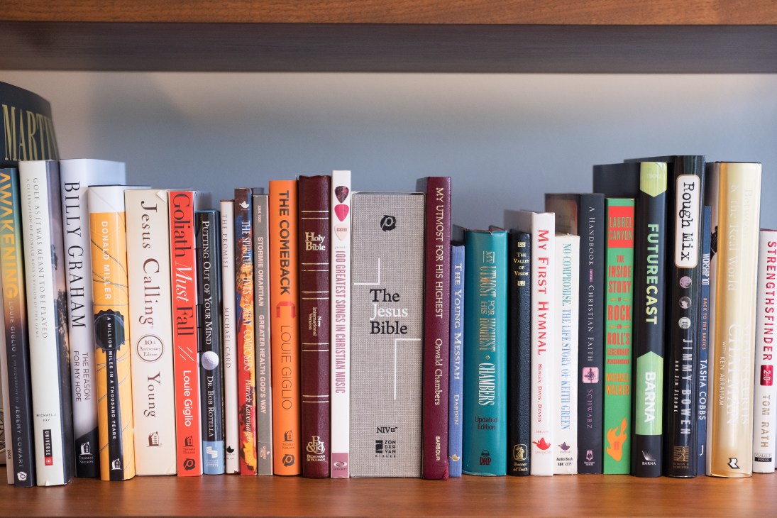 A collection of books and Bibles standing upright on a wooden shelf, including titles about faith, Christianity and spirituality.