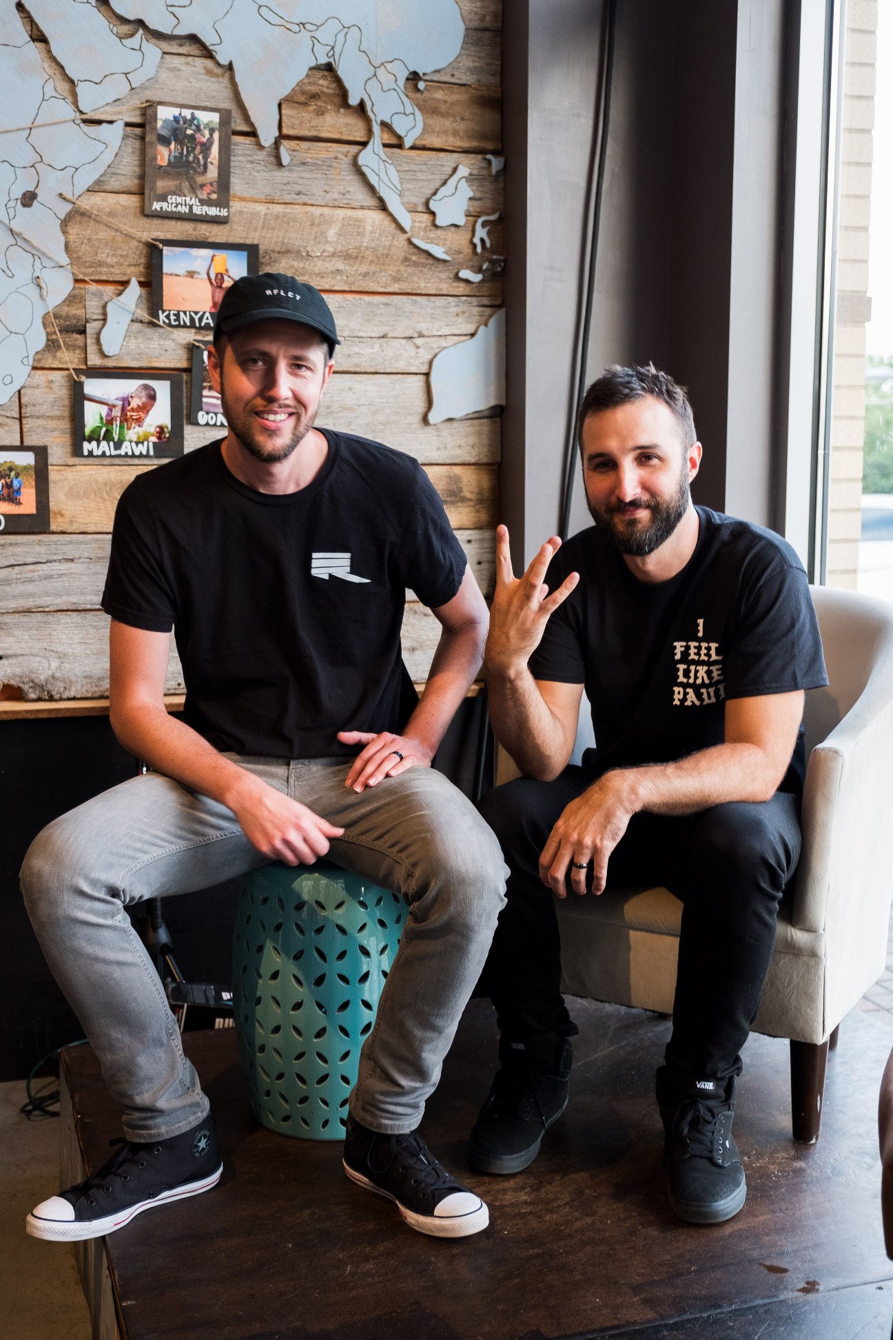 Two men seated indoors against a wooden wall with travel photographs, both wearing black t-shirts.