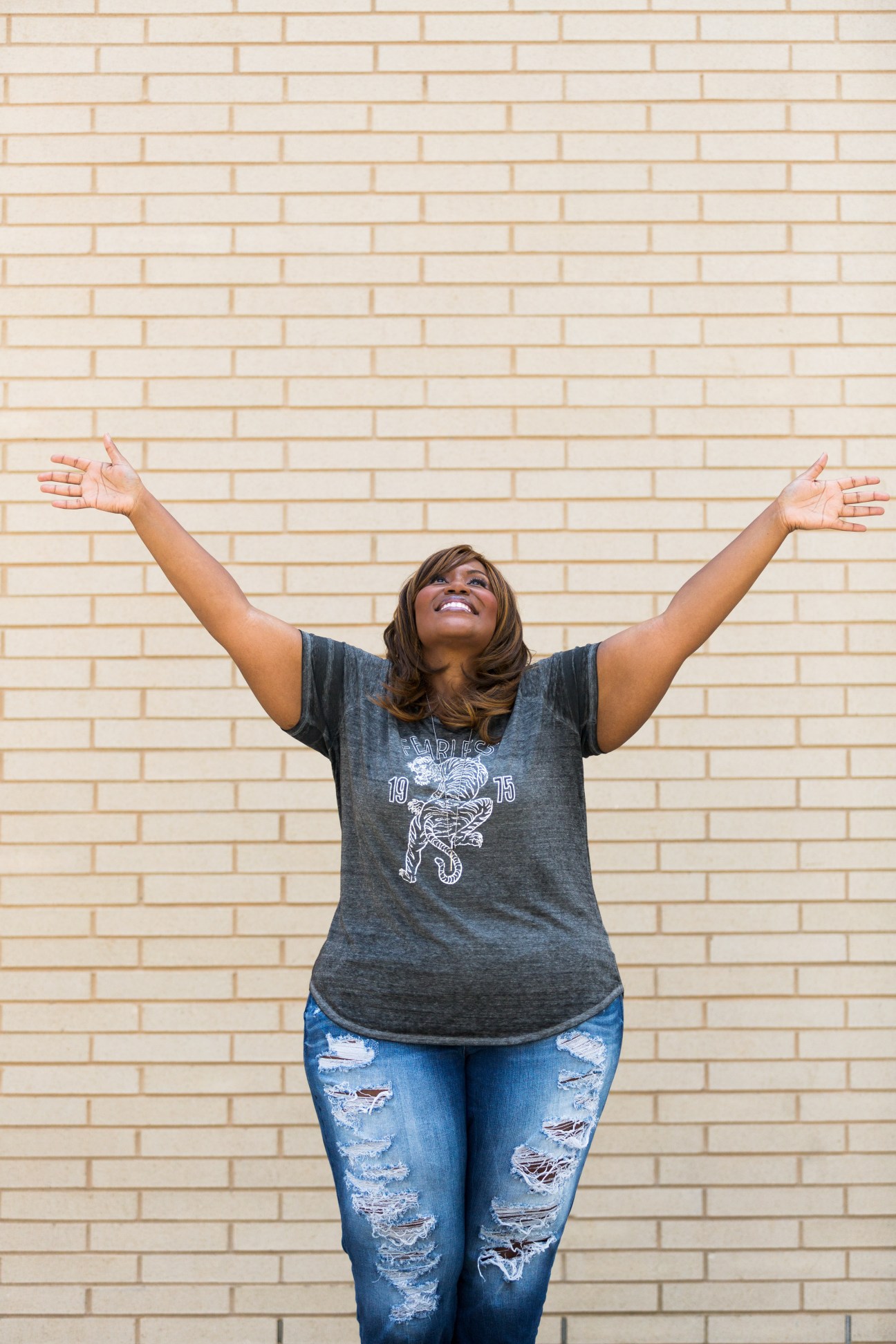 Singer Mandisa standing with arms raised against a brick wall, wearing a gray graphic t-shirt and distressed blue jeans.