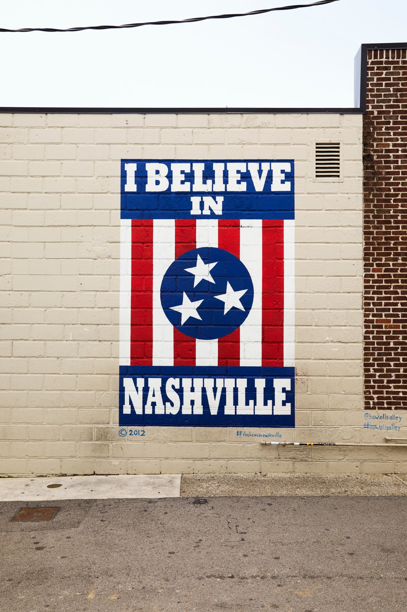 Street mural reading 'I BELIEVE IN NASHVILLE' with red and white stripes and blue circle with stars on cream brick wall.