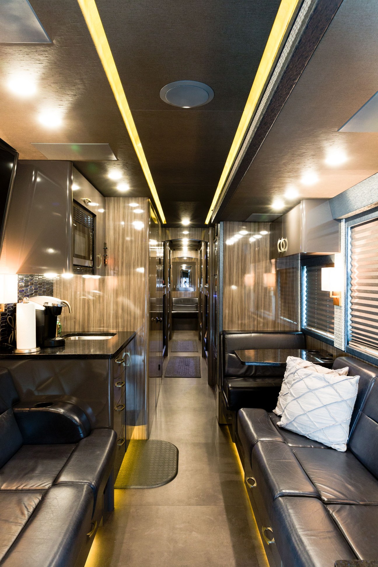 Interior of a luxury tour bus with black leather seating, ambient yellow lighting, and a kitchenette area.