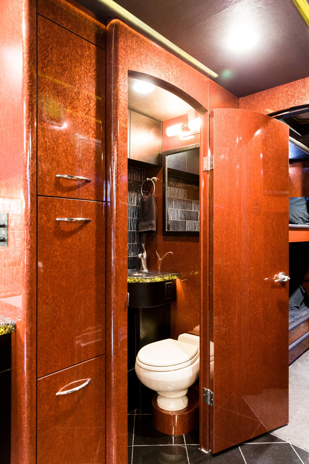 Compact bathroom with rust-coloured walls, white toilet, black sink with gold accents, and recessed lighting in a tour bus.