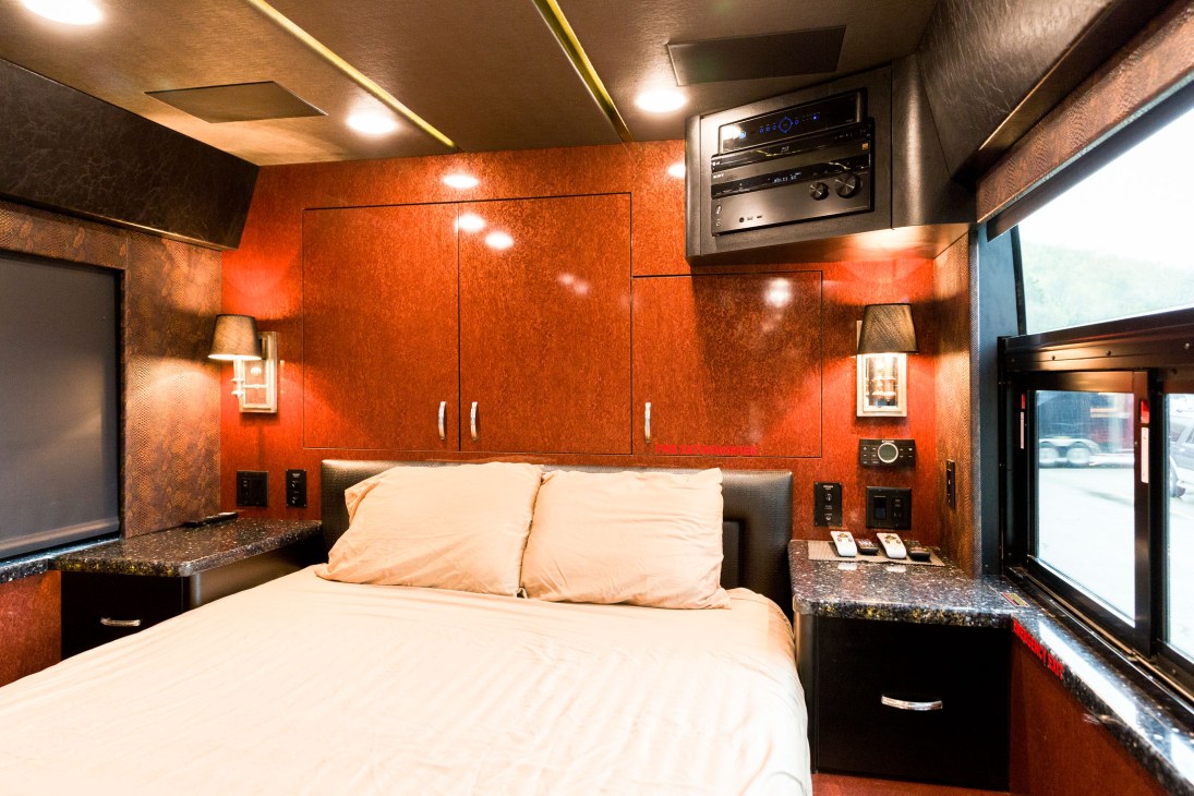 Luxury tour bus bedroom with orange-red wood paneling, queen bed, built-in cabinets, granite countertops and windows.