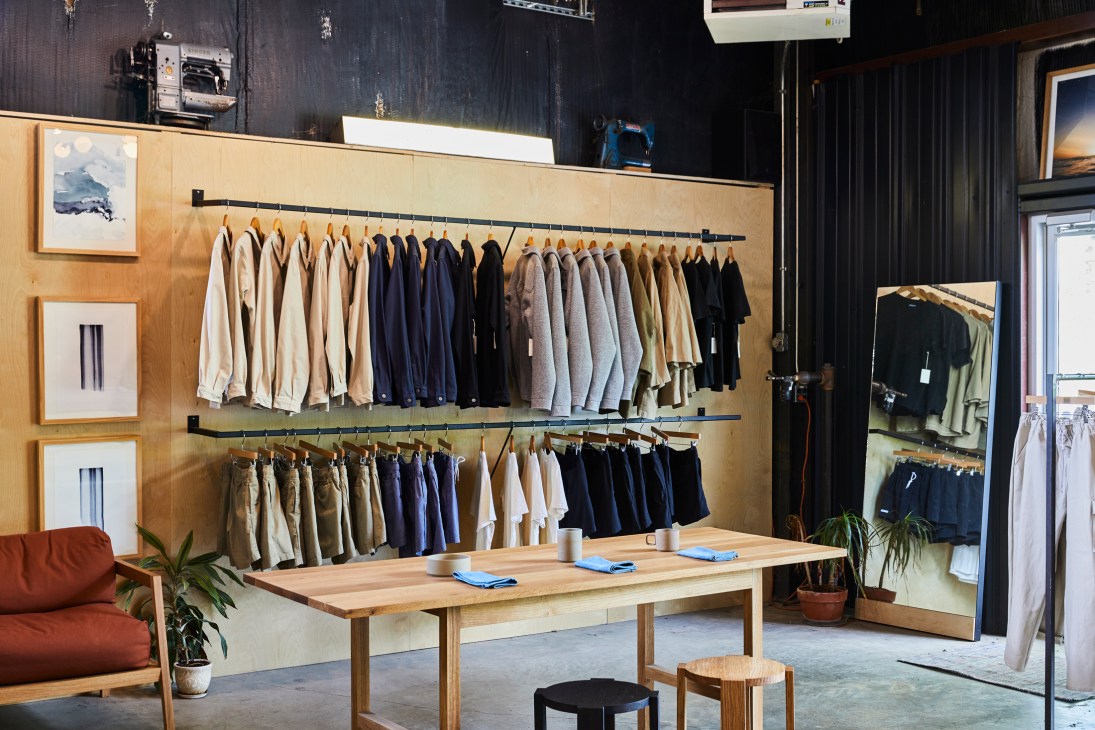 Oil + Lumber showroom with wooden wall displaying rows of neutral and navy clothing on black rods, wooden table with blue items, and full-length mirror.