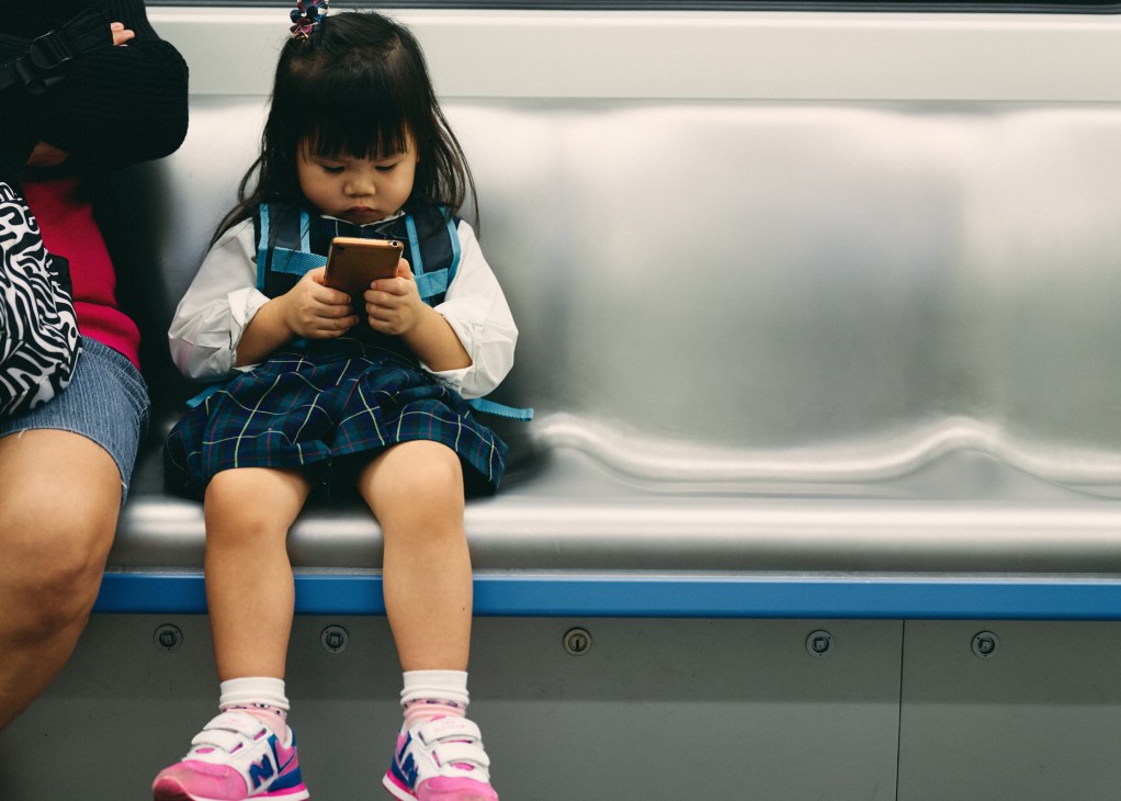 Young child sitting on a subway train seat holding a mobile device.