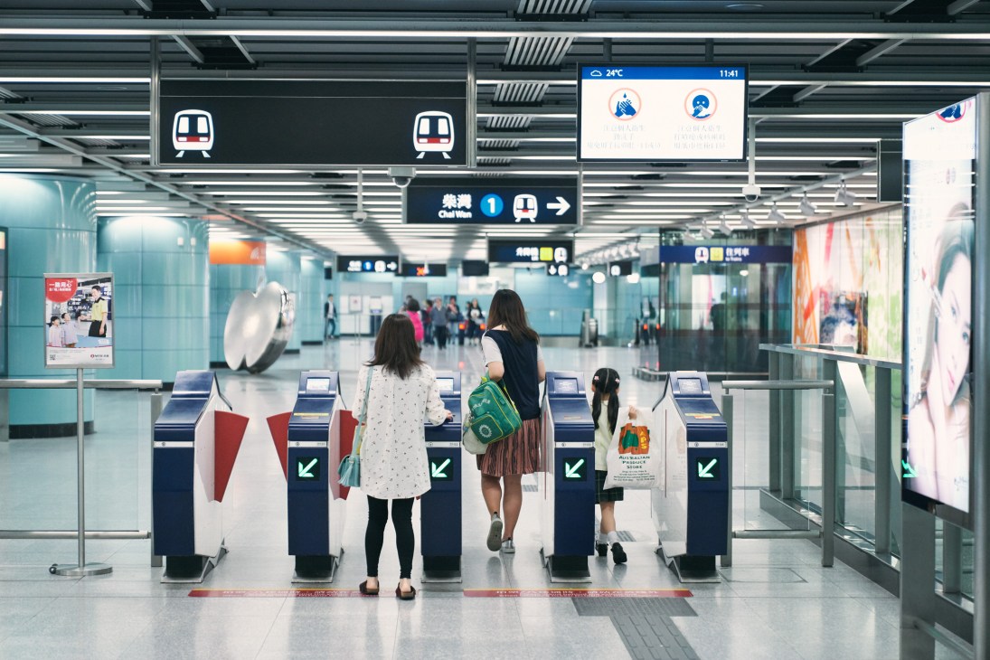 Commuters pass through a Hong Kong MTR station with directional signage and ticket gates overhead.