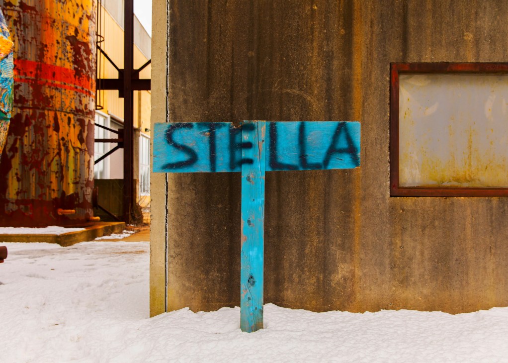 Weathered industrial exterior with blue wooden sign reading 'STELLA' against rust-stained metal walls.