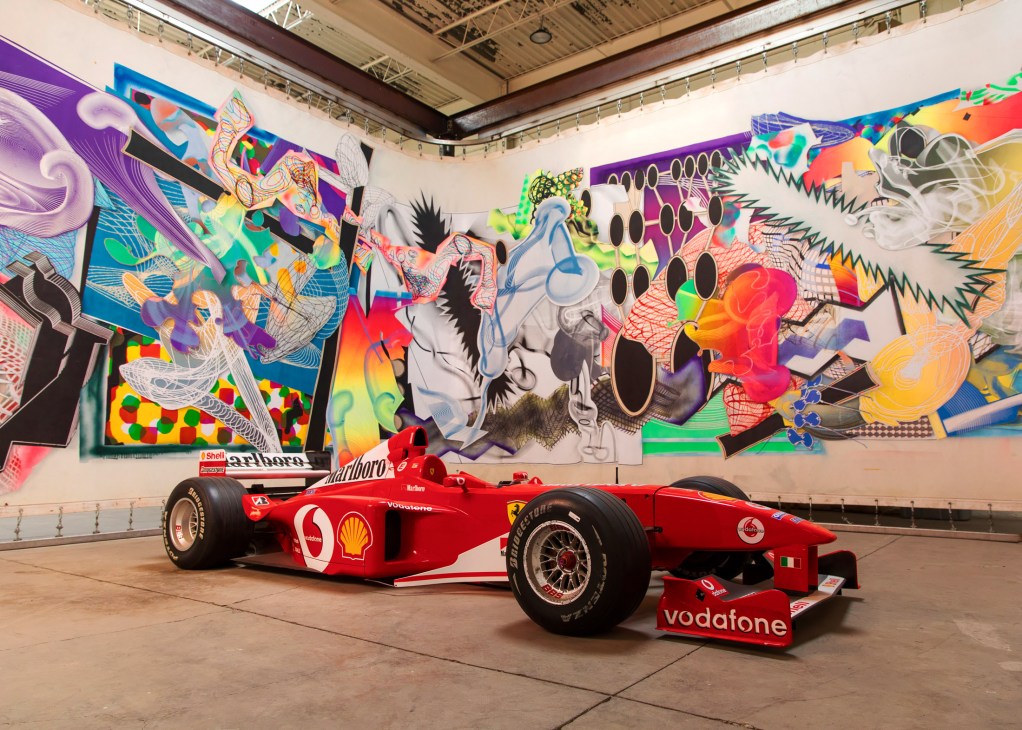 Red Formula 1 car displayed beneath a large colorful abstract mural with vibrant patterns and shapes.