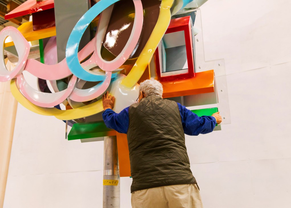 Frank Stella examining one of his colorful abstract sculptures featuring bright pink, blue, yellow, and orange curved forms.