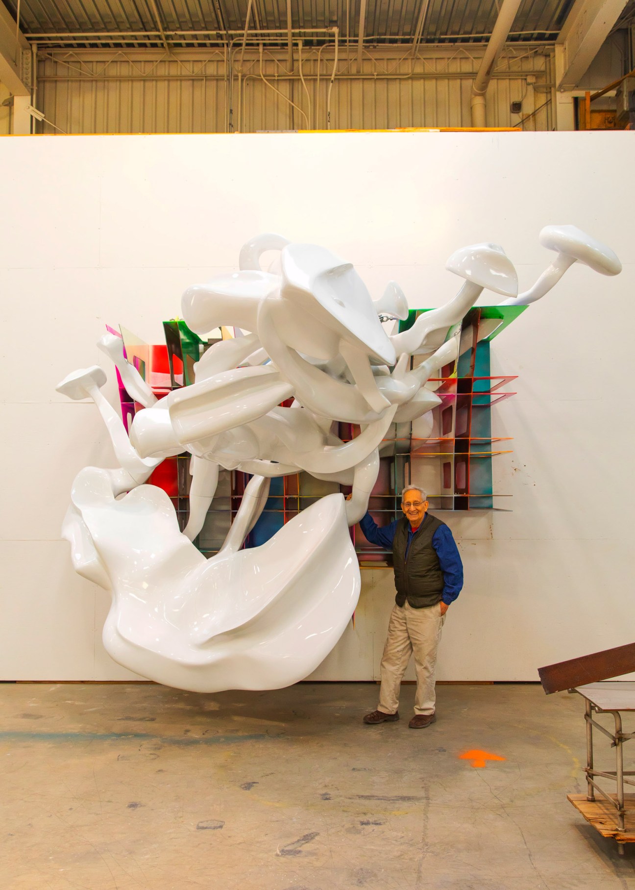 Frank Stella stands beside a large white abstract sculpture with colorful geometric elements in his studio.