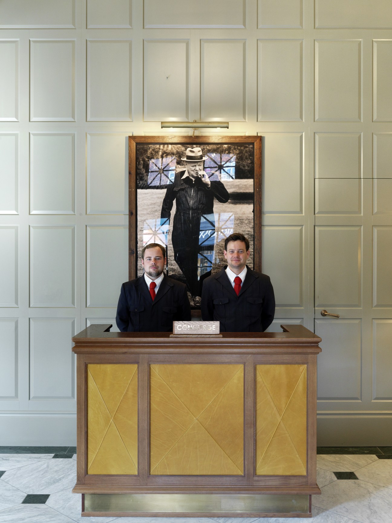 Two concierge staff in dark suits with red ties stand behind a wooden desk with gold panels beneath a framed vintage photograph.
