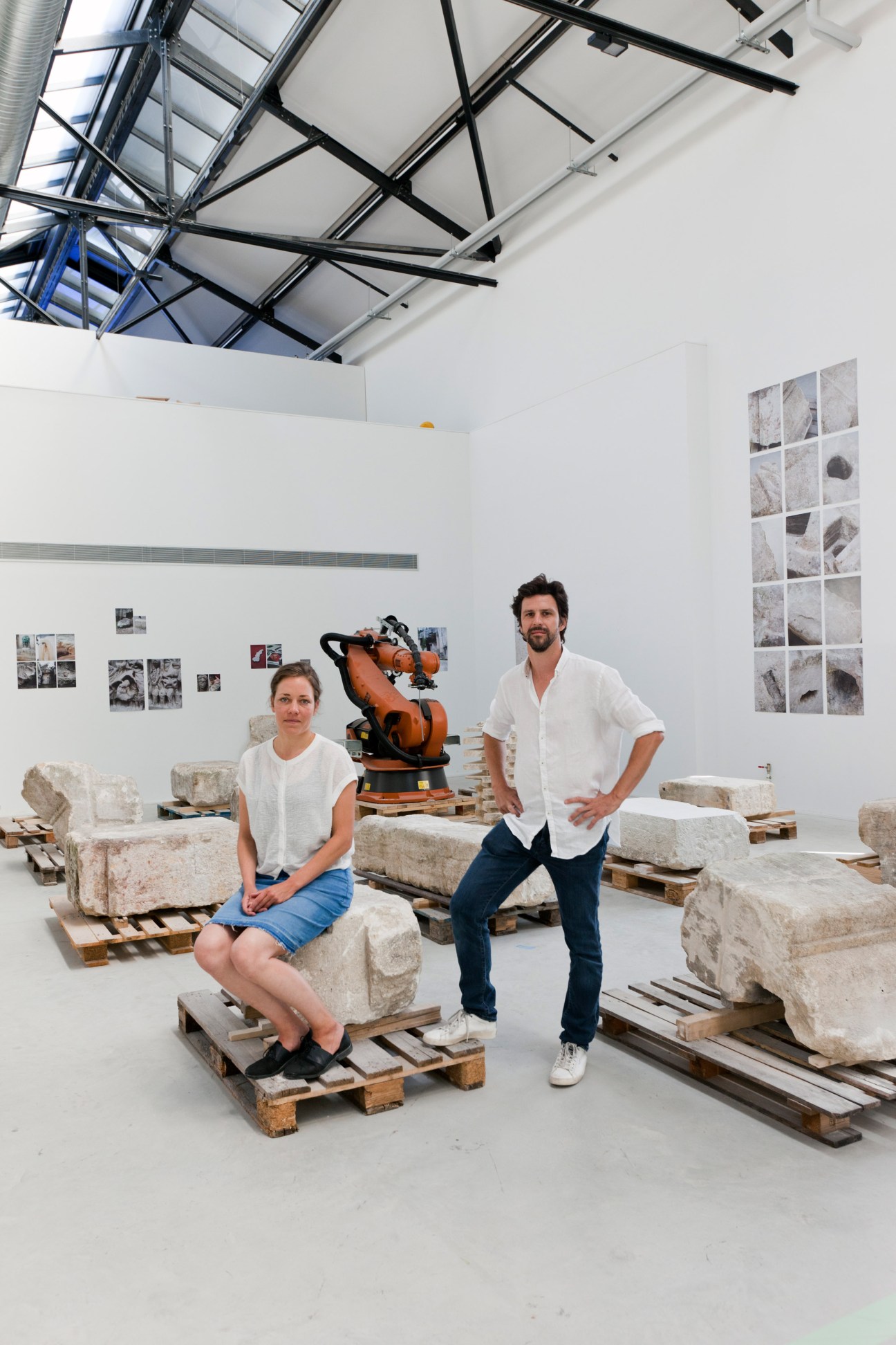 Two people in white shirts standing among stone blocks on wooden pallets in a modern gallery space