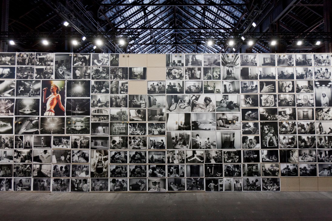 Large wall display of hundreds of black and white photographs arranged in a grid pattern in industrial gallery space