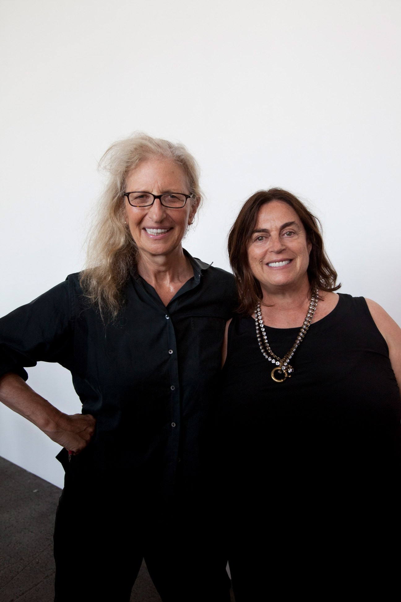 Two women standing together smiling, one with long blonde hair and glasses, one with dark hair wearing a necklace